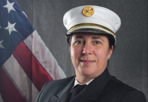 Photo of Kassandra Whidden in Class A uniform in front of draped American Flag